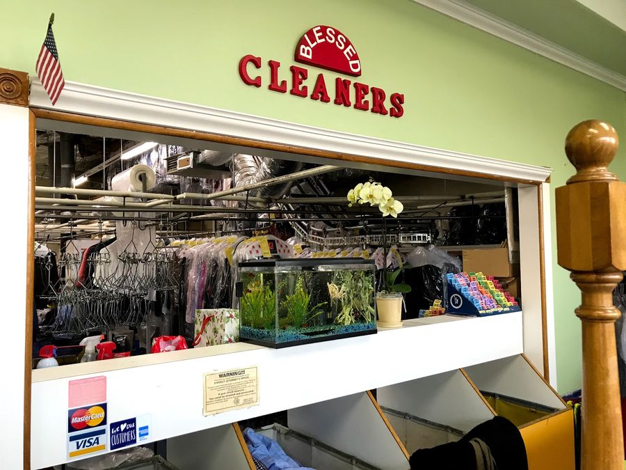 The Blessed Cleaners front counter: a red arched “Blessed Cleaners” sign above the pass-through window, with bagged garments on the conveyor behind, an orchid and a small aquarium on the ledge.