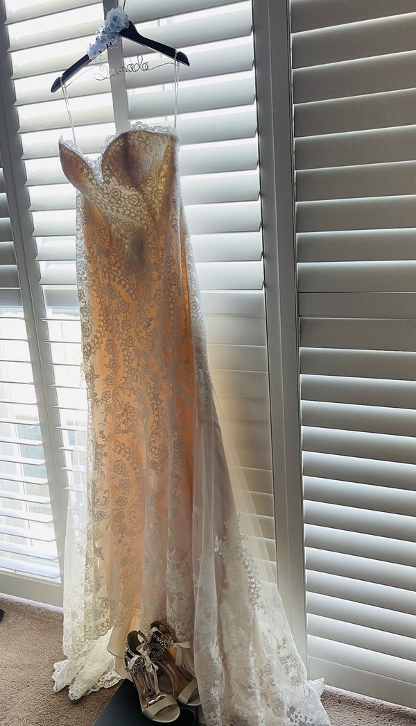 A lace wedding gown hanging on a hanger in front of white window shutters.