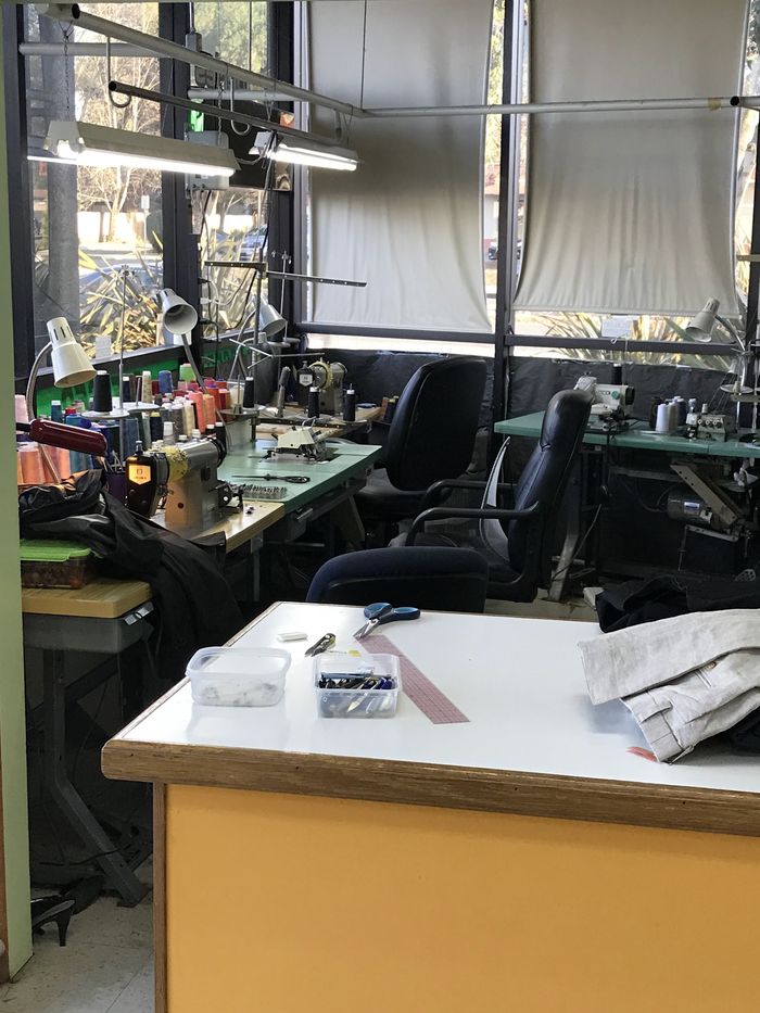 The alterations workroom: industrial sewing machines with spools of thread along a bright window, and a cutting table in front.